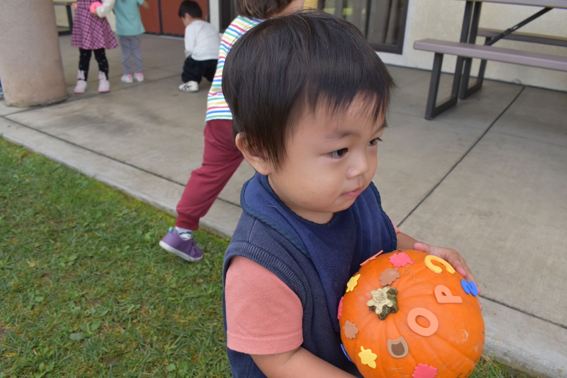 Child holding a pumpkin decorated with letters outdoors, other children play nearby.