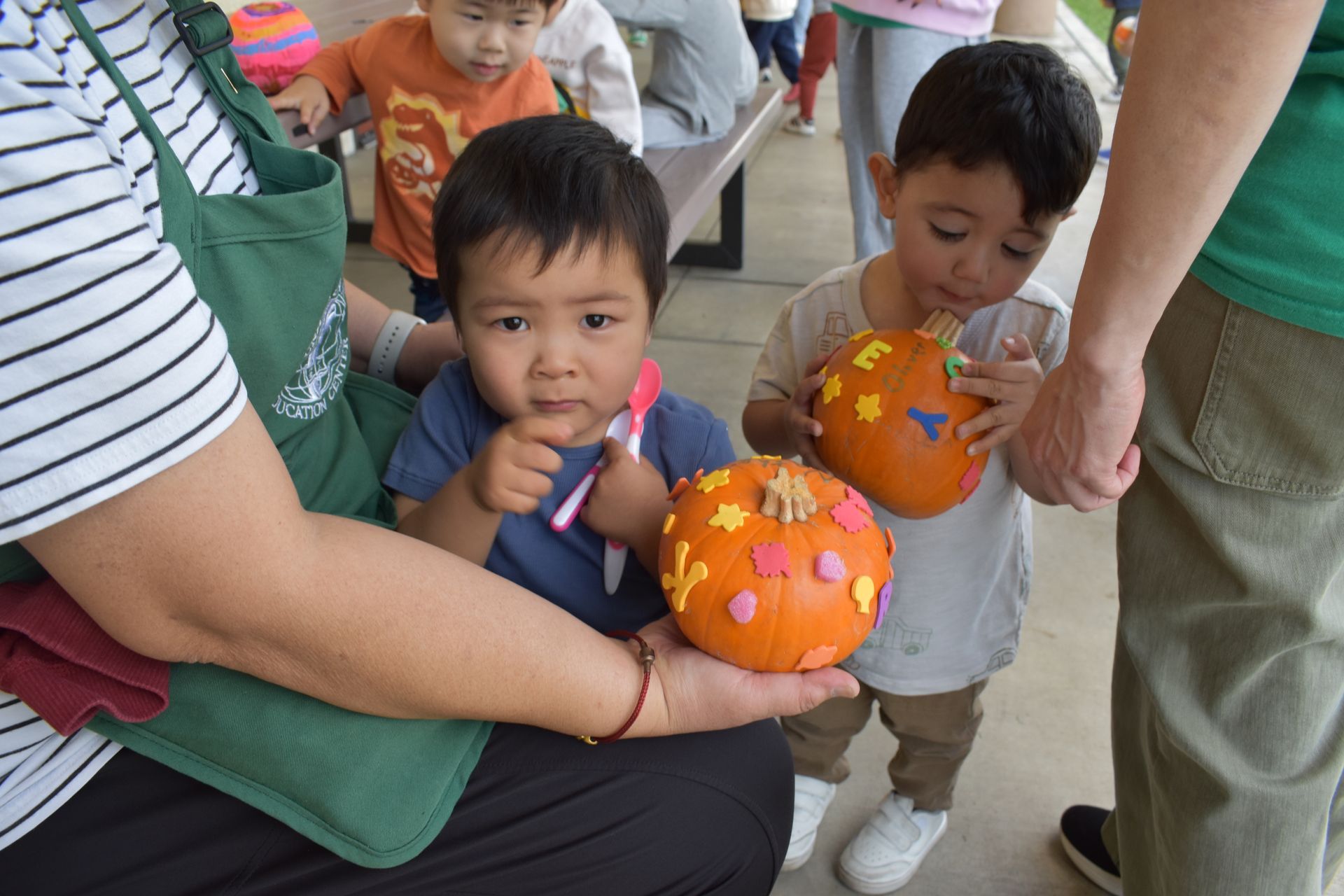 Two children with decorated pumpkins held by adults at an outdoor event.