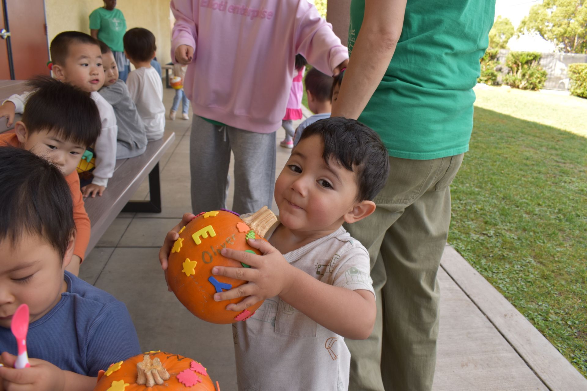 Child holding a decorated pumpkin smiles, others in background. Outdoors, sunny.