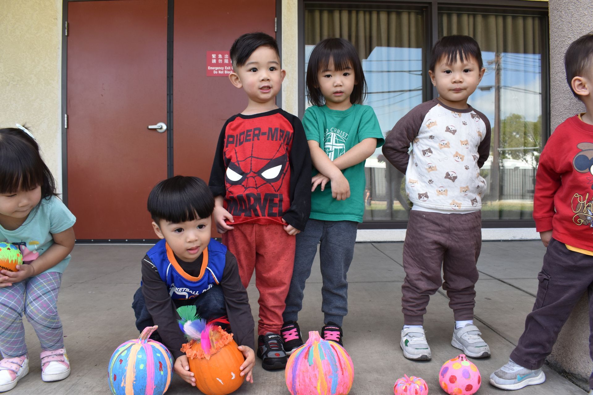 Children standing and kneeling with painted pumpkins outside a building.