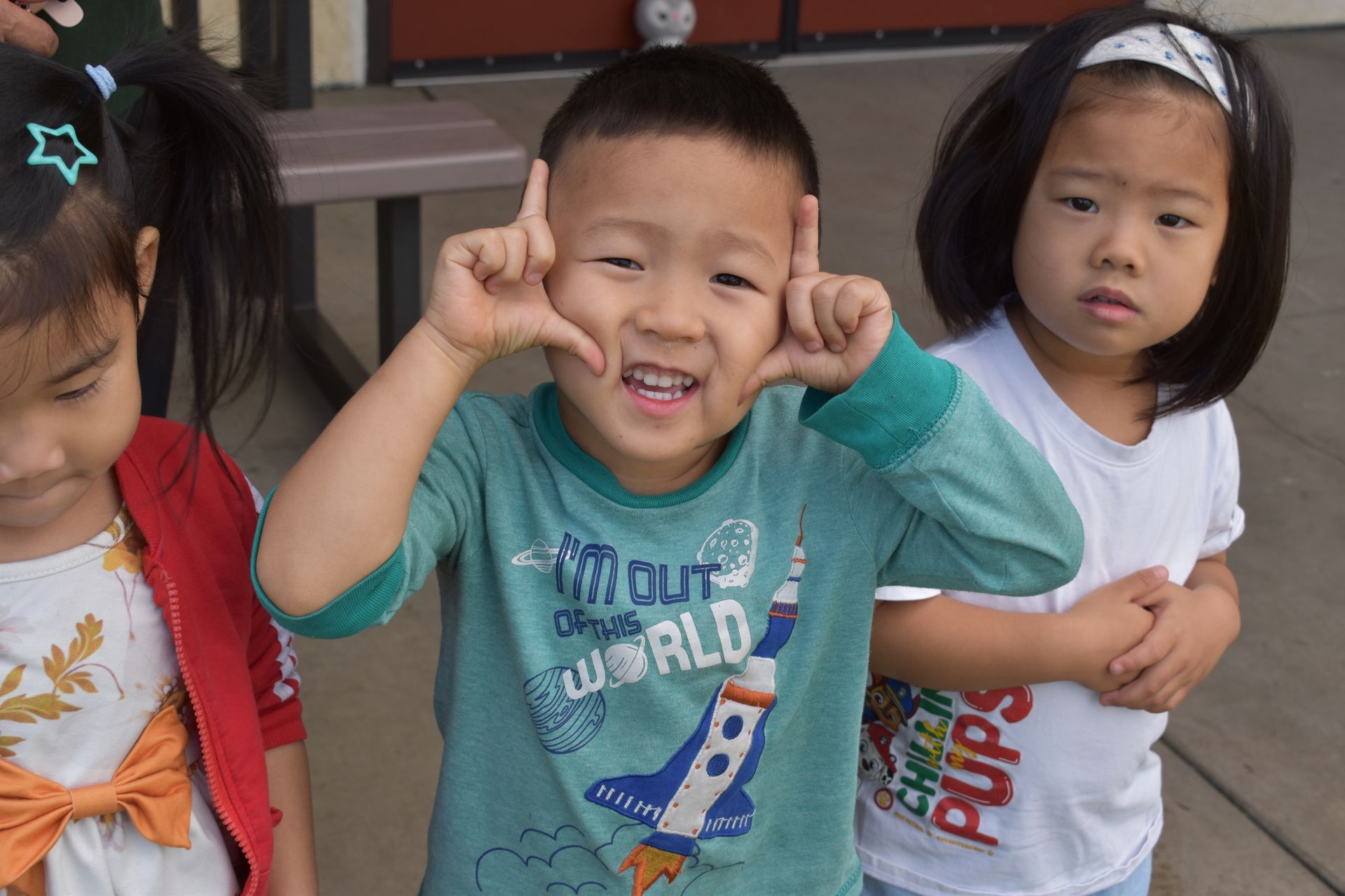 Three children posing outdoors; boy making frame with fingers, smiling; girls looking at camera.