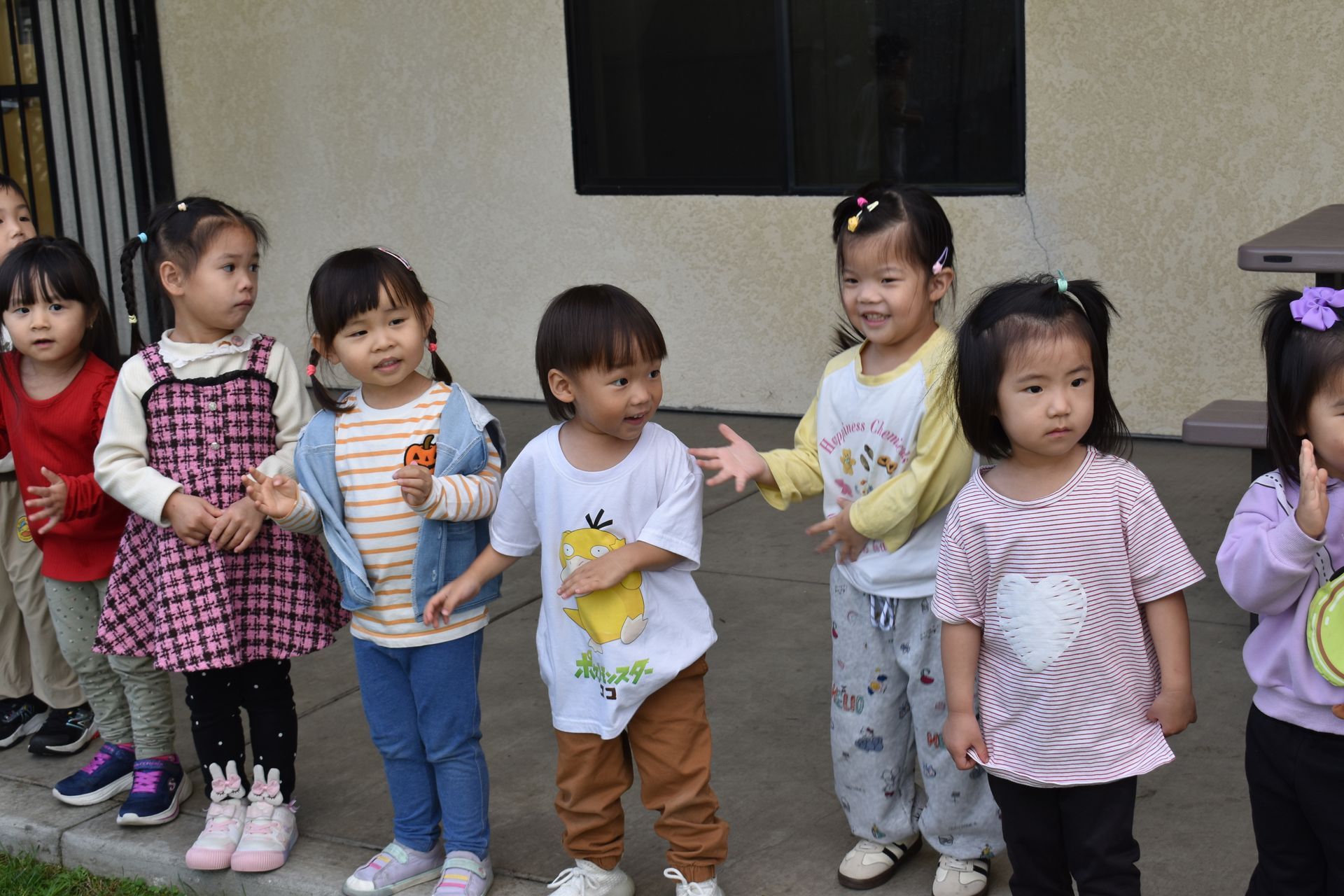 Group of young children outdoors, standing in a row, some smiling.