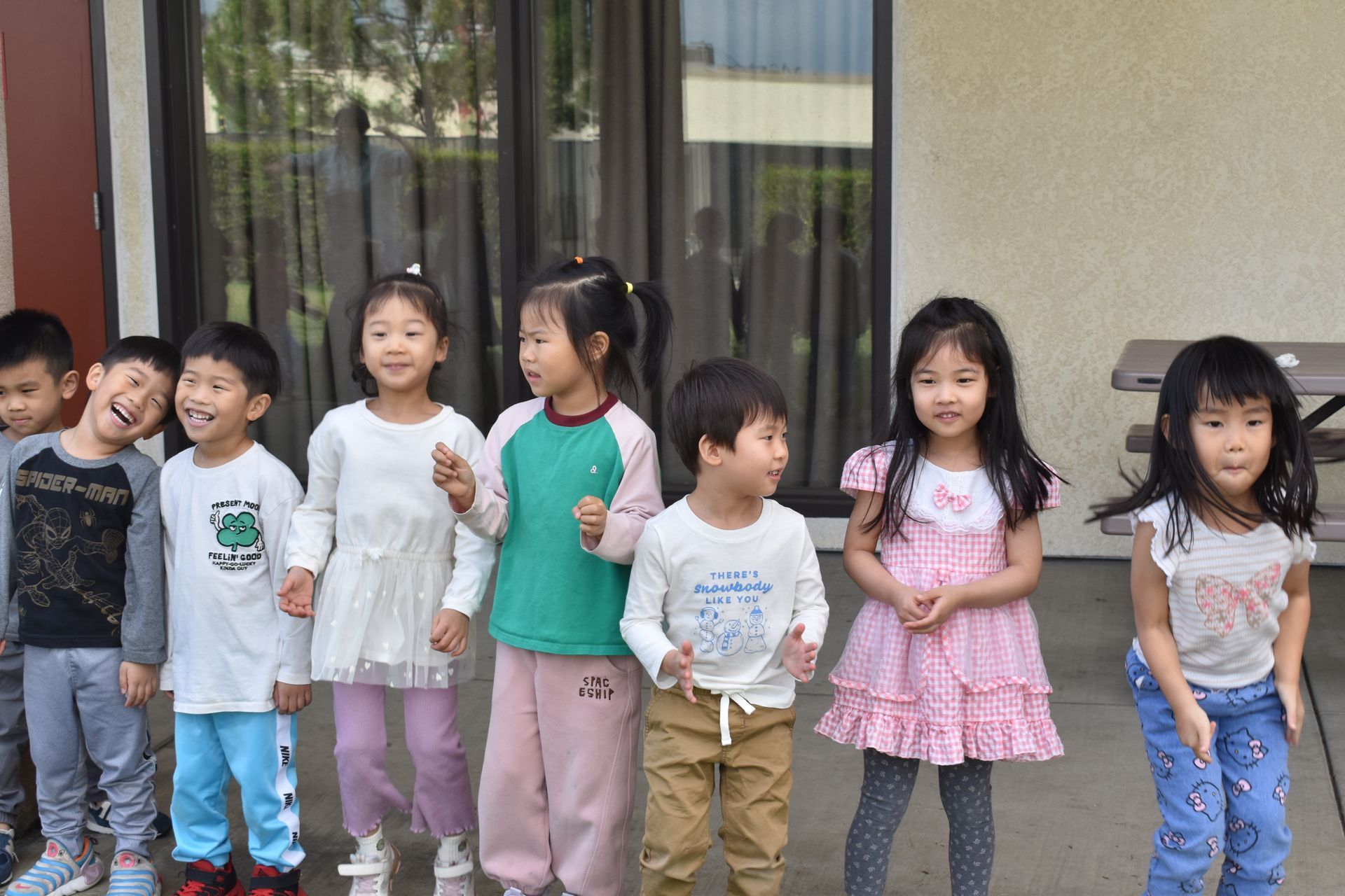 Group of children standing outside near windows, some smiling.
