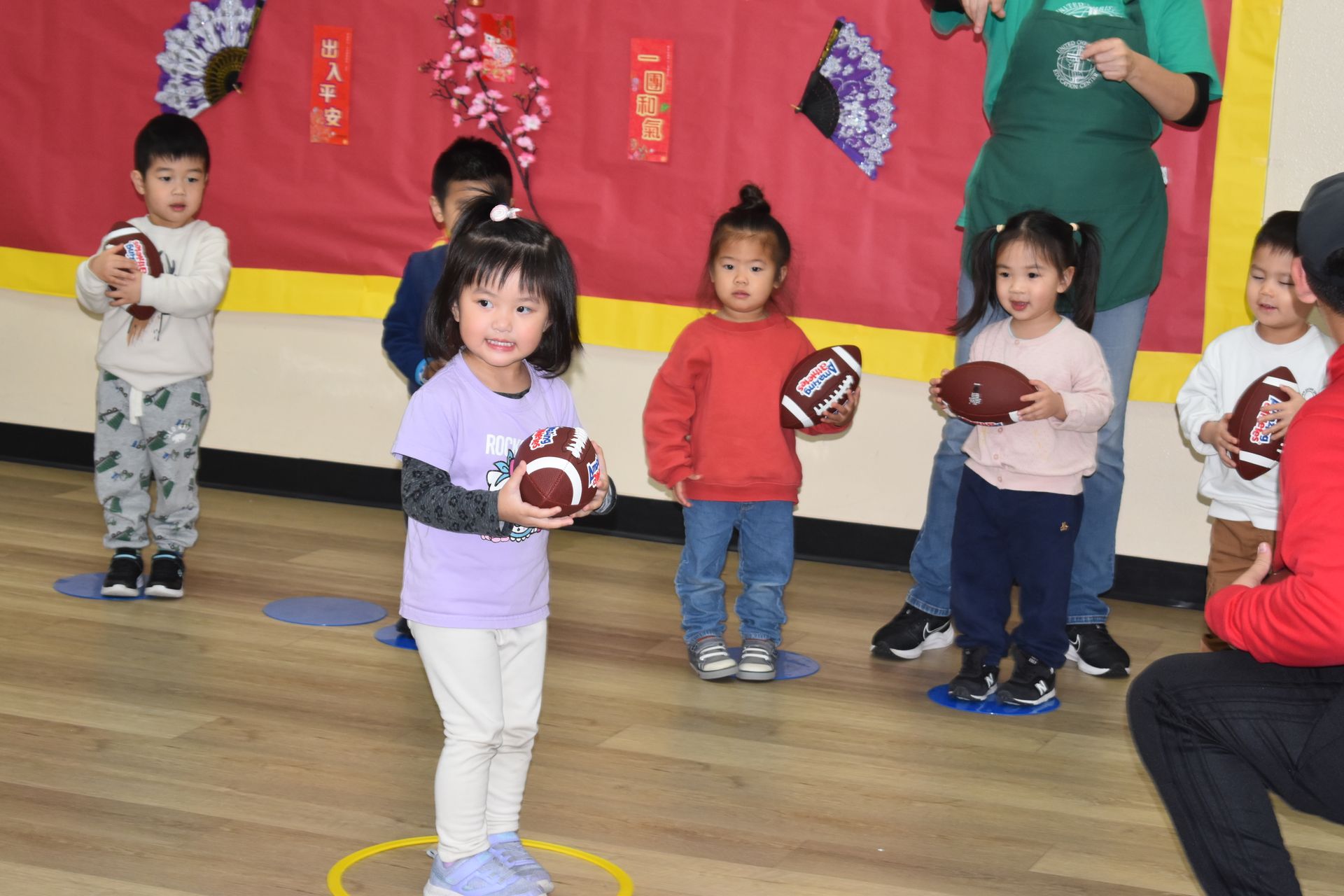 Children holding footballs in a classroom; adults present.