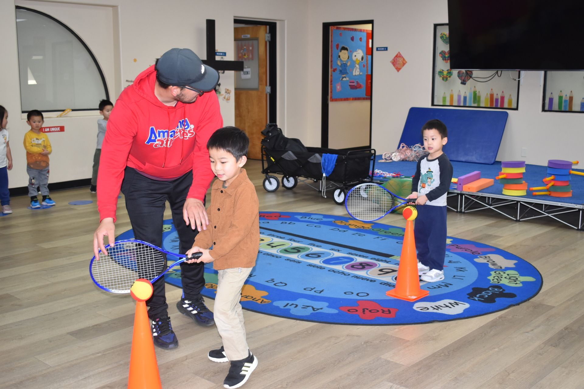 Man helping a child practice tennis indoors with rackets, balls, and cones on a blue rug. Other children watch.