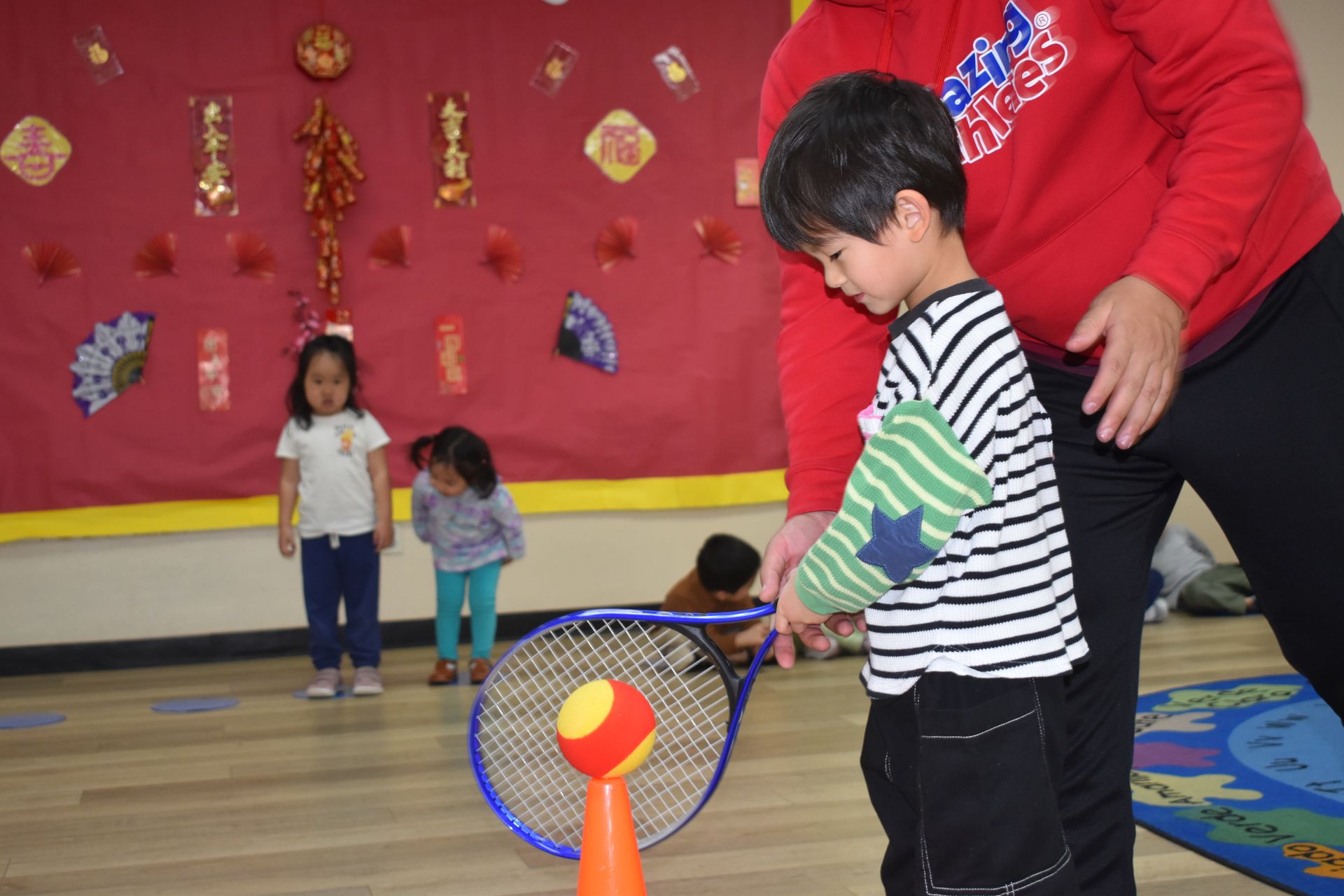 Boy aiming ball with racquet at cone, supervised by an adult indoors near other children and Chinese New Year decorations.