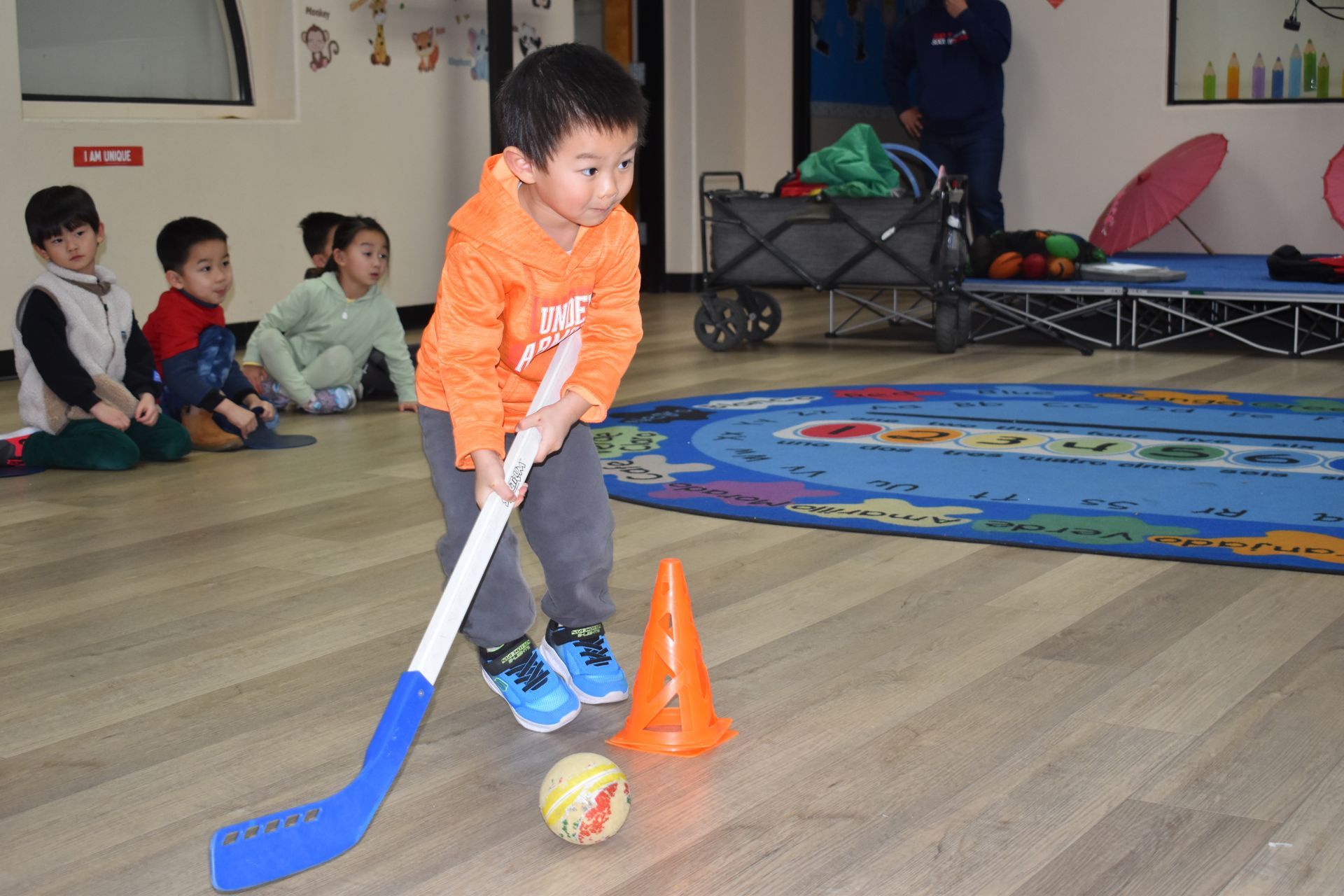 A child playing hockey indoors, aiming stick at ball, orange cone, other children watching.