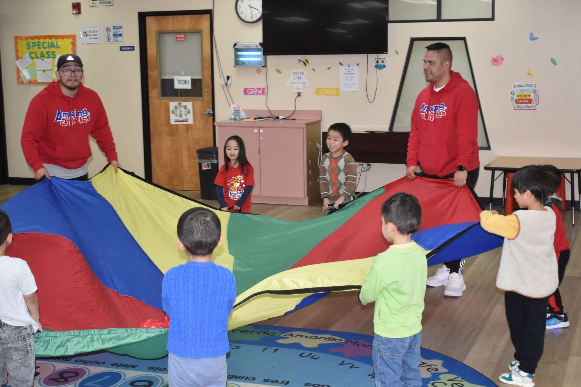 Children and two adults holding a rainbow parachute indoors.