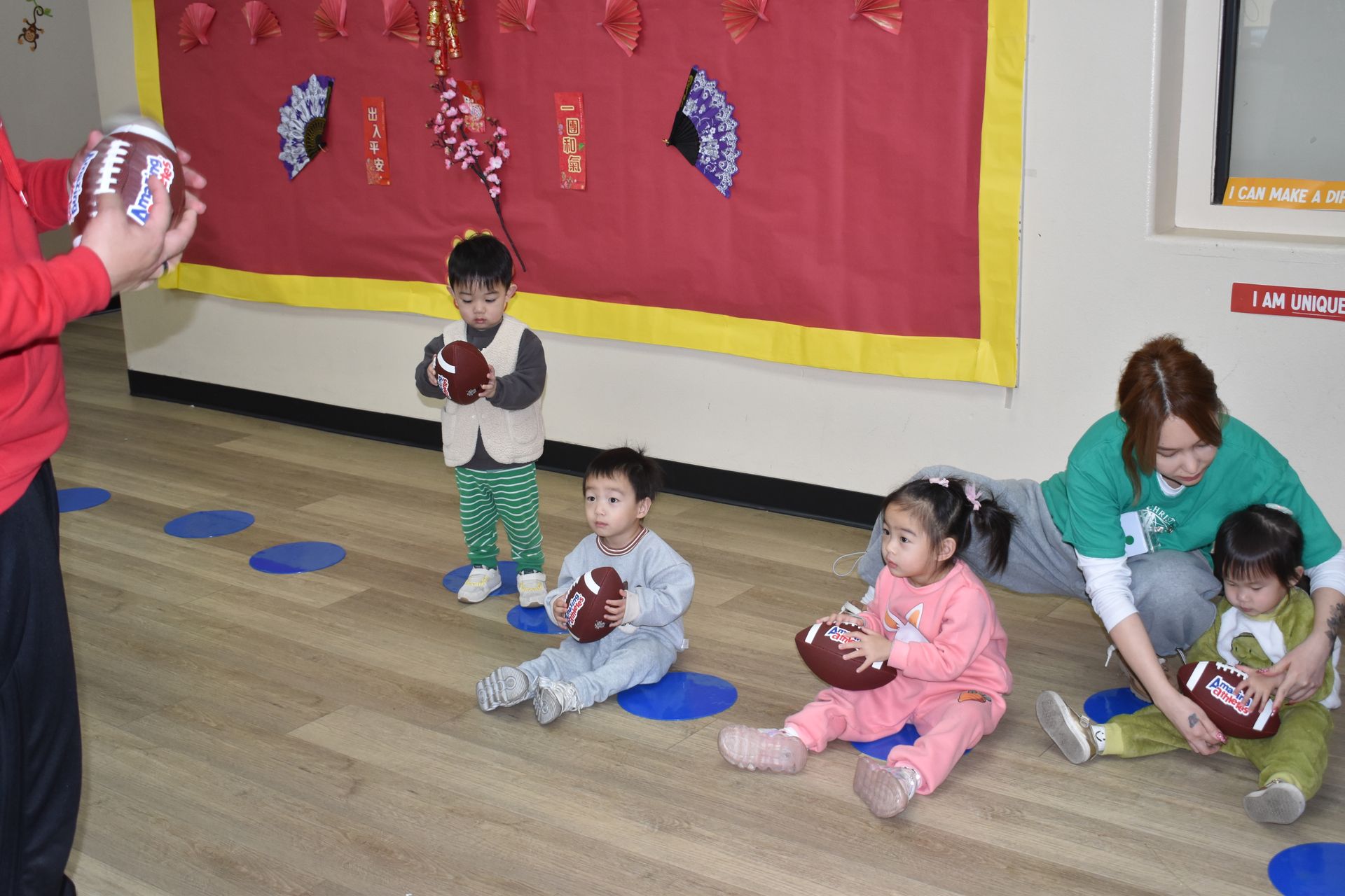 Children in a playroom holding footballs, guided by adults. Red wall decor.