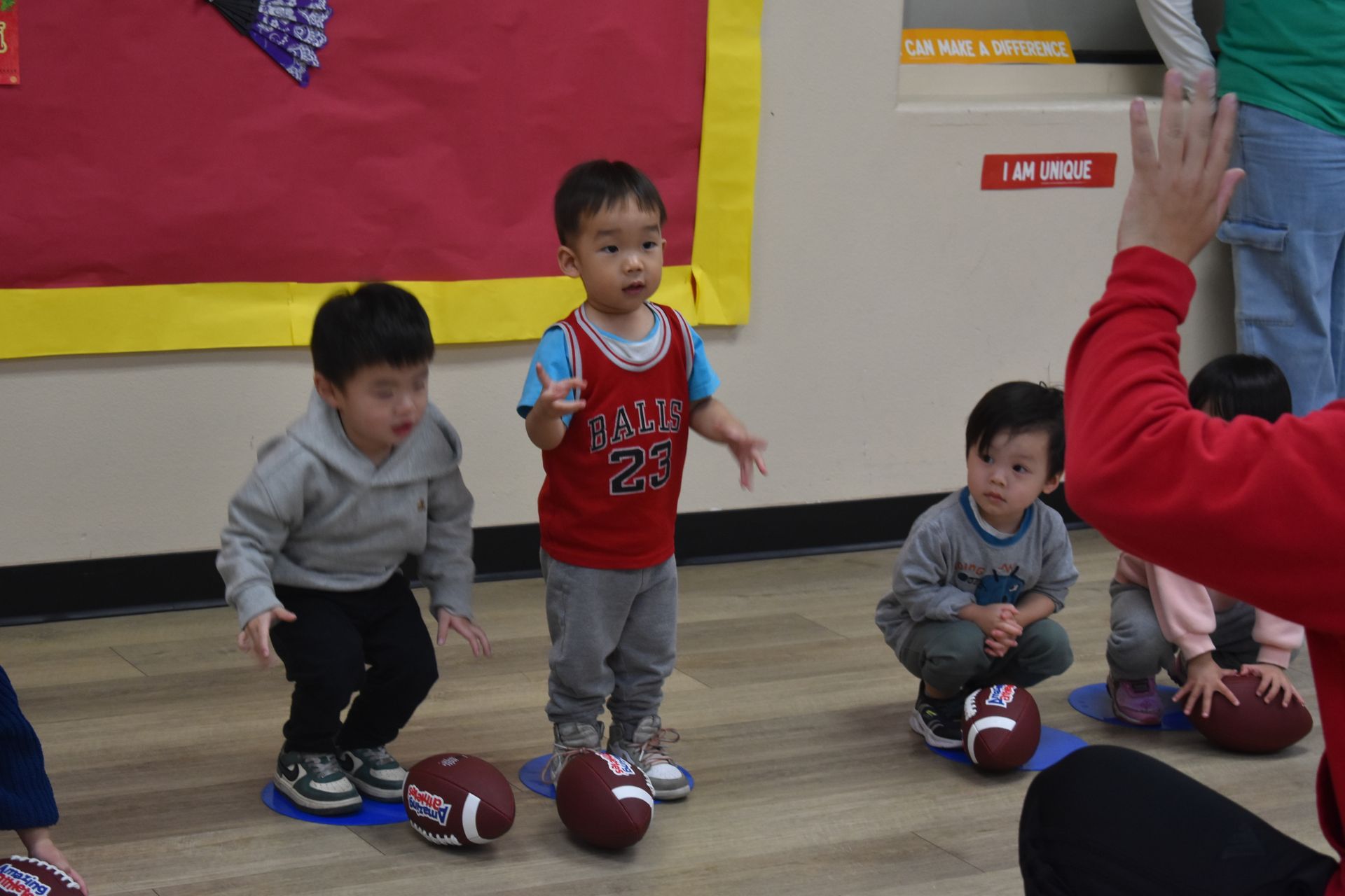 Children standing and squatting with footballs in a classroom; a person's hand raised.