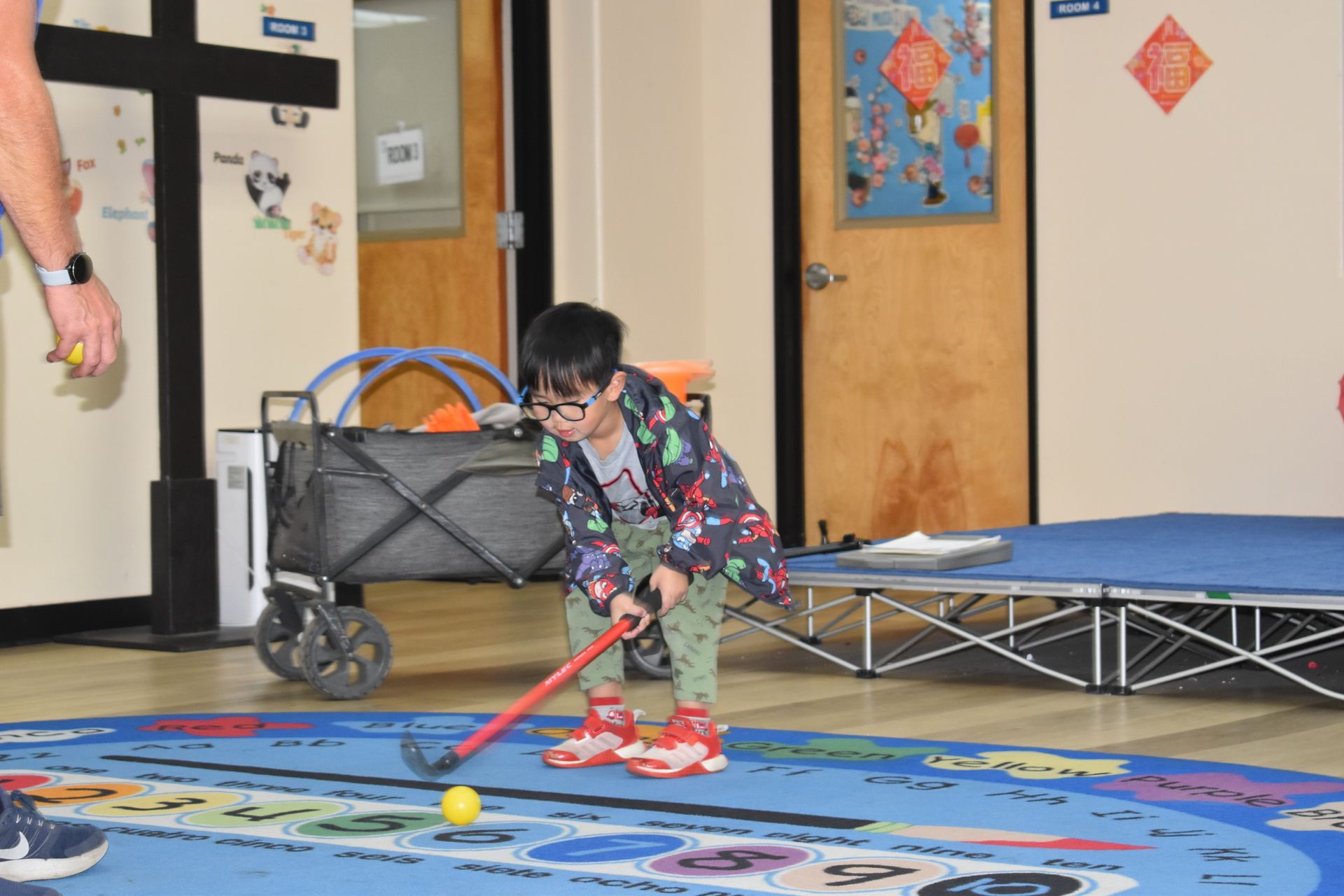 Child playing floor hockey indoors.  Yellow ball, red hockey stick, carpeted floor.  Cart in the background.