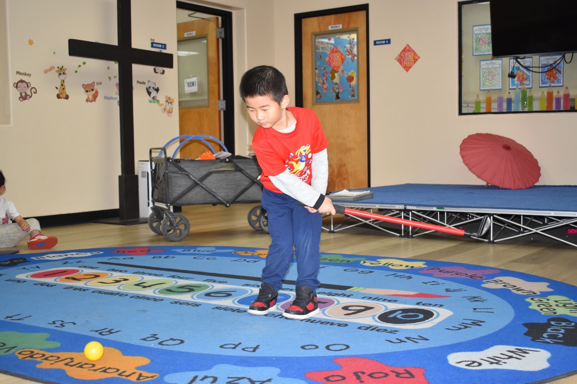 Boy in red shirt swings a toy golf club on a colorful rug inside a room.