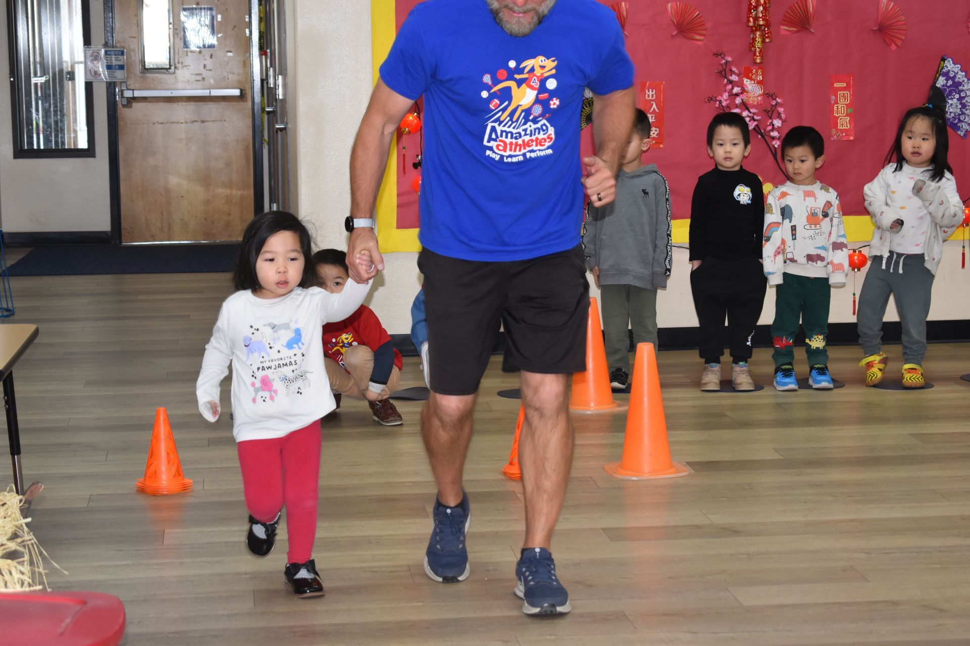 Man and child run together indoors, holding hands; children watch. Orange cones on floor.