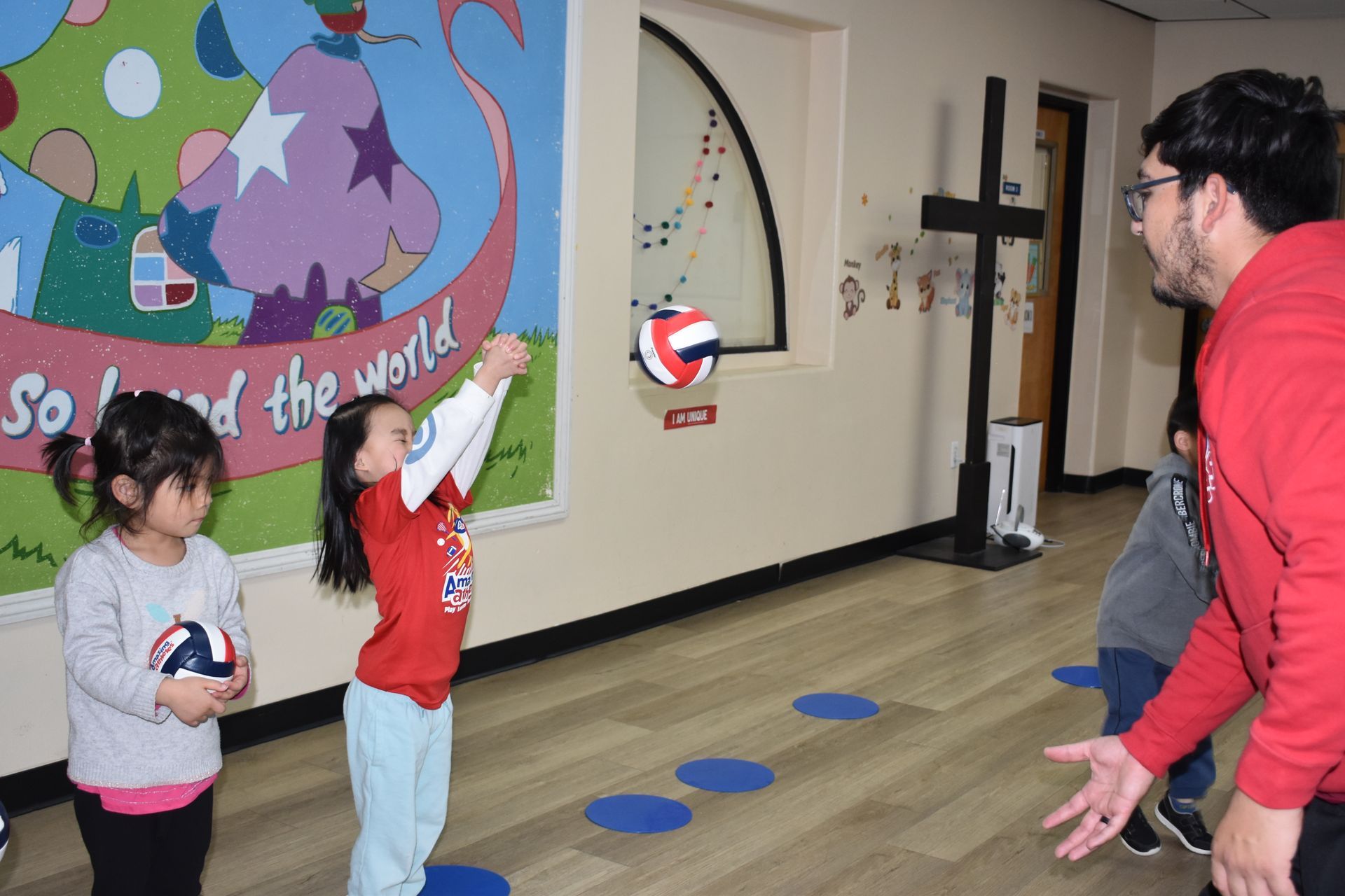 A man in red coaches a child catching a volleyball. Another child watches. Indoor setting.