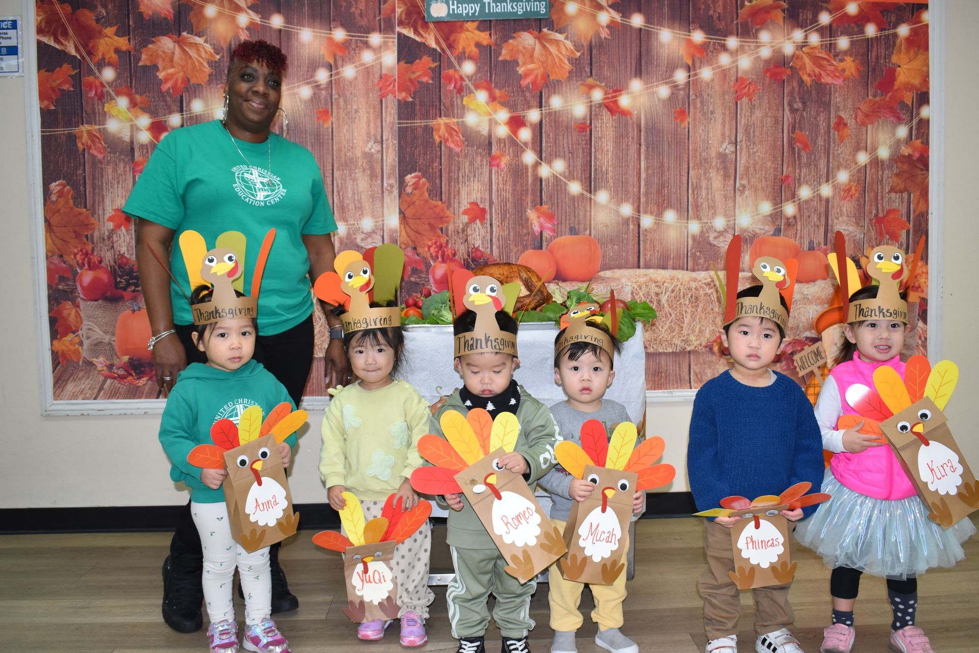 A teacher and children pose with turkey crafts in front of a fall-themed backdrop.