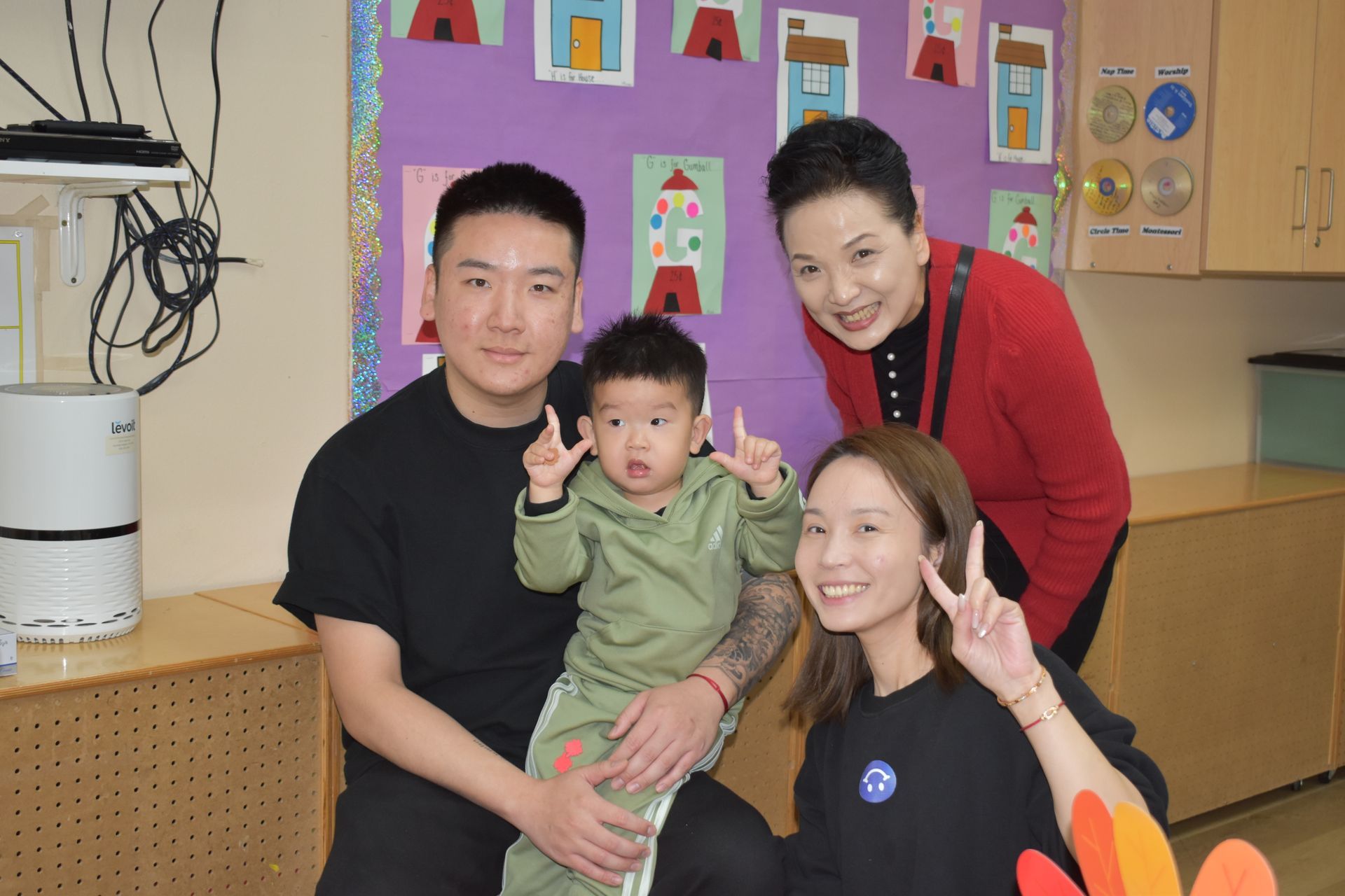 Family of four poses indoors. A man, woman, and child sit, while another woman stands behind them smiling.