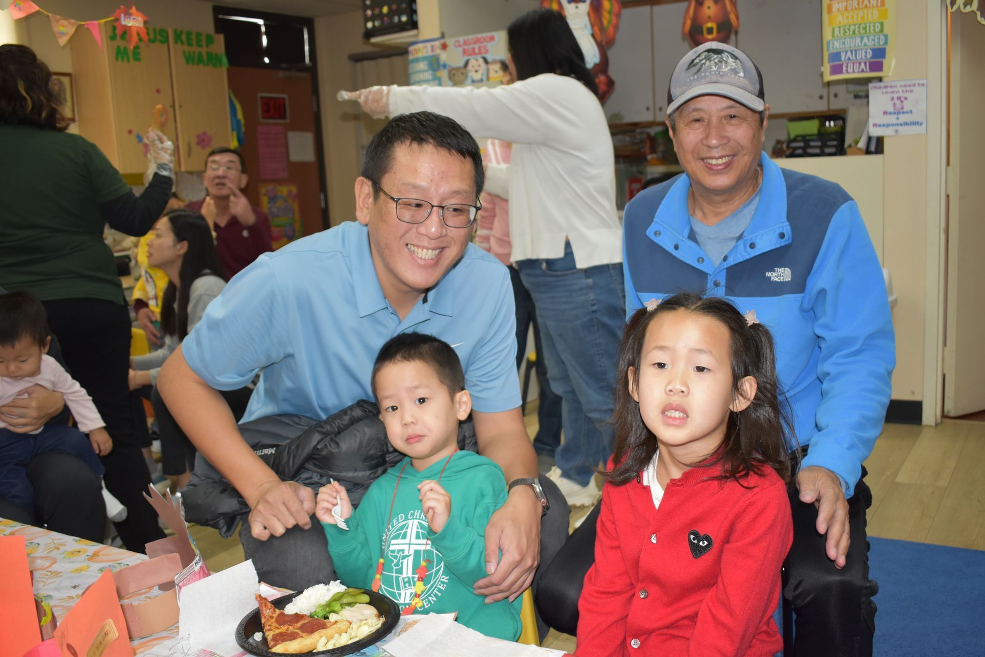 Two men and two children smiling at a table indoors; one man has a blue jacket, the other a blue polo.