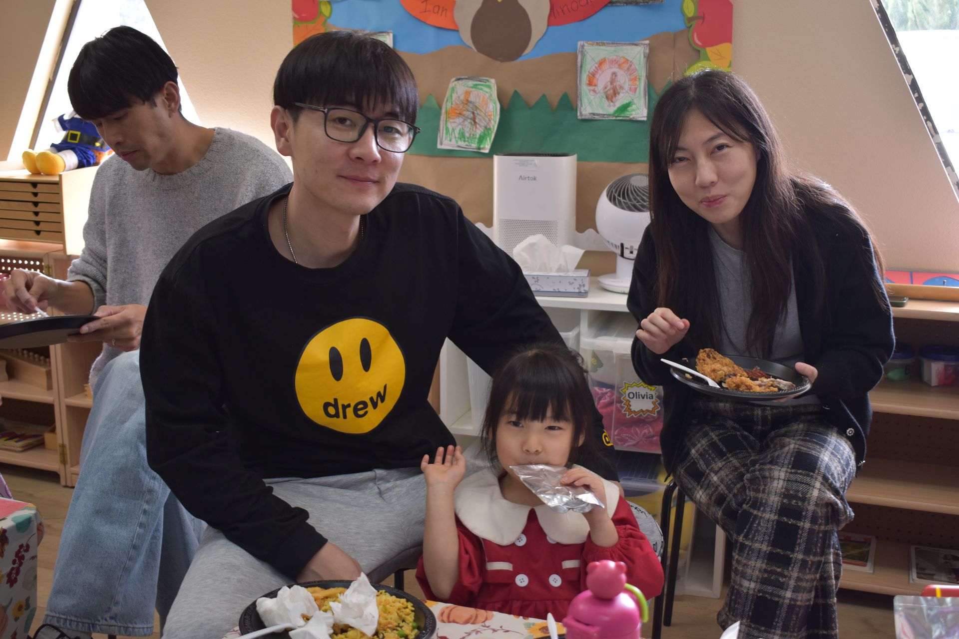 Family enjoying food at a table indoors; man, woman, child, and another person visible.