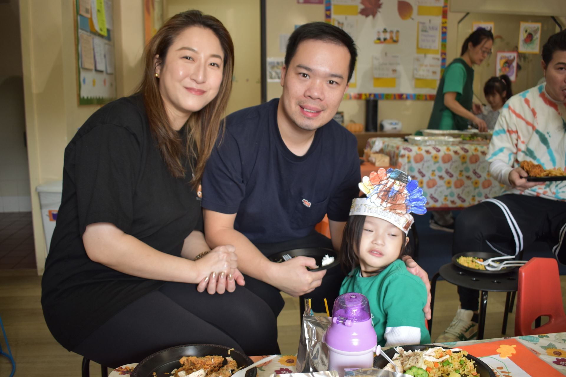 Family in a classroom setting, smiling with a child wearing a paper hat. Food on table.