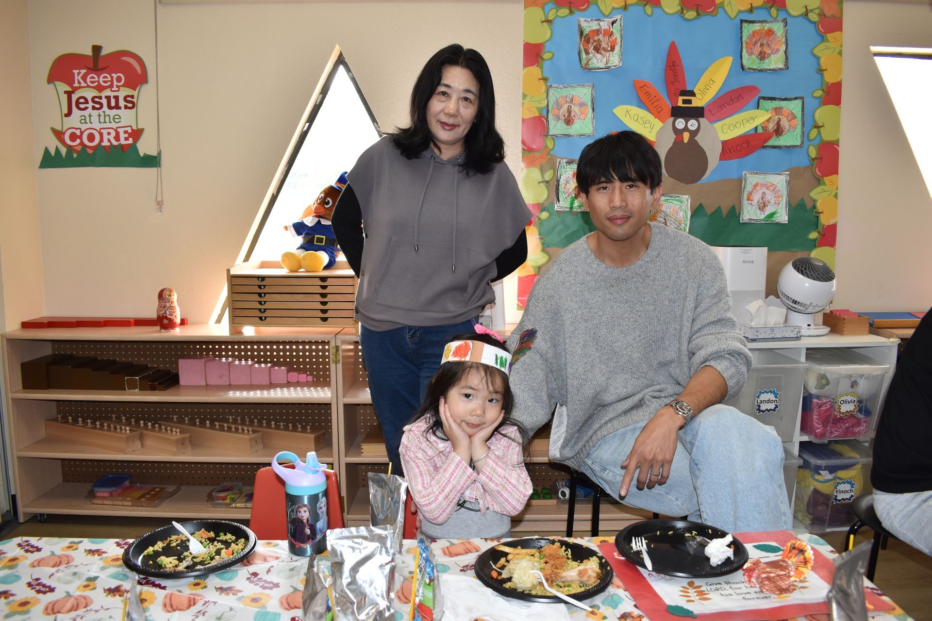 Family at a table with food, child wearing a headband, woman standing, man seated, room with decorations.