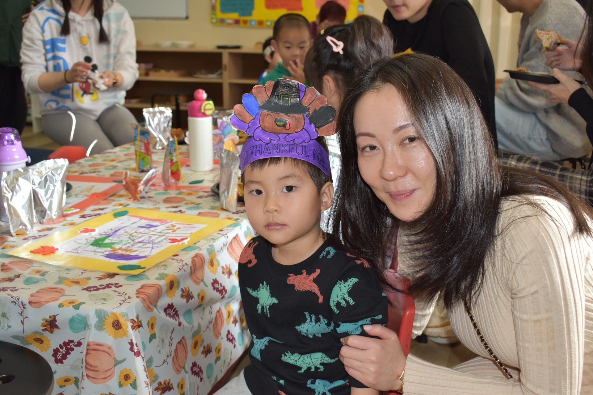 A young child and an adult smile at the camera. The child wears a turkey hat. They sit at a craft table.