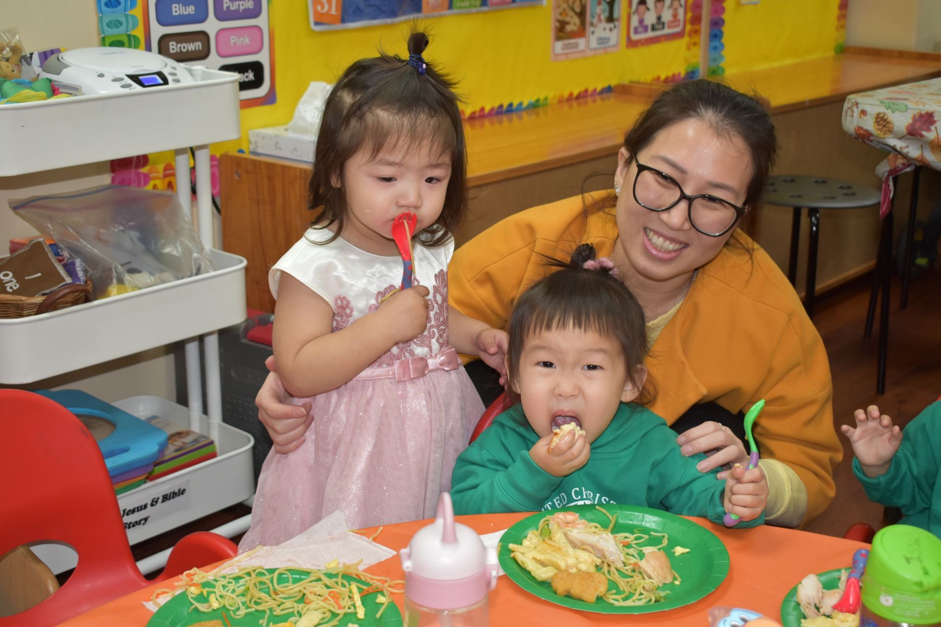 A woman and two young children eating at a table. The woman smiles, children use spoons, food on plates.