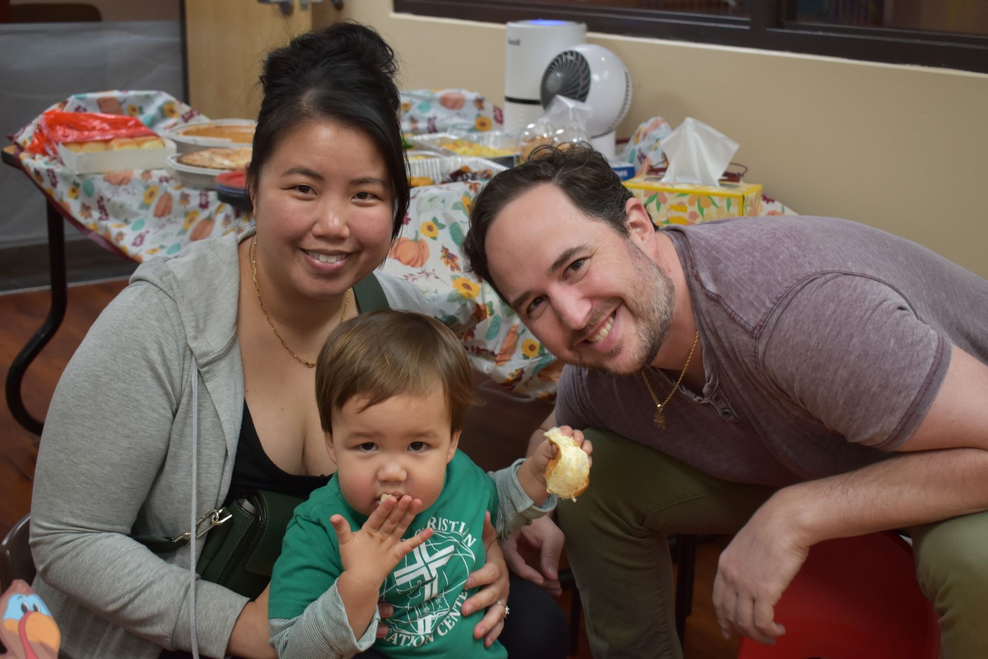 A family of three smiles for the camera. The child eats food, while the adults sit nearby.