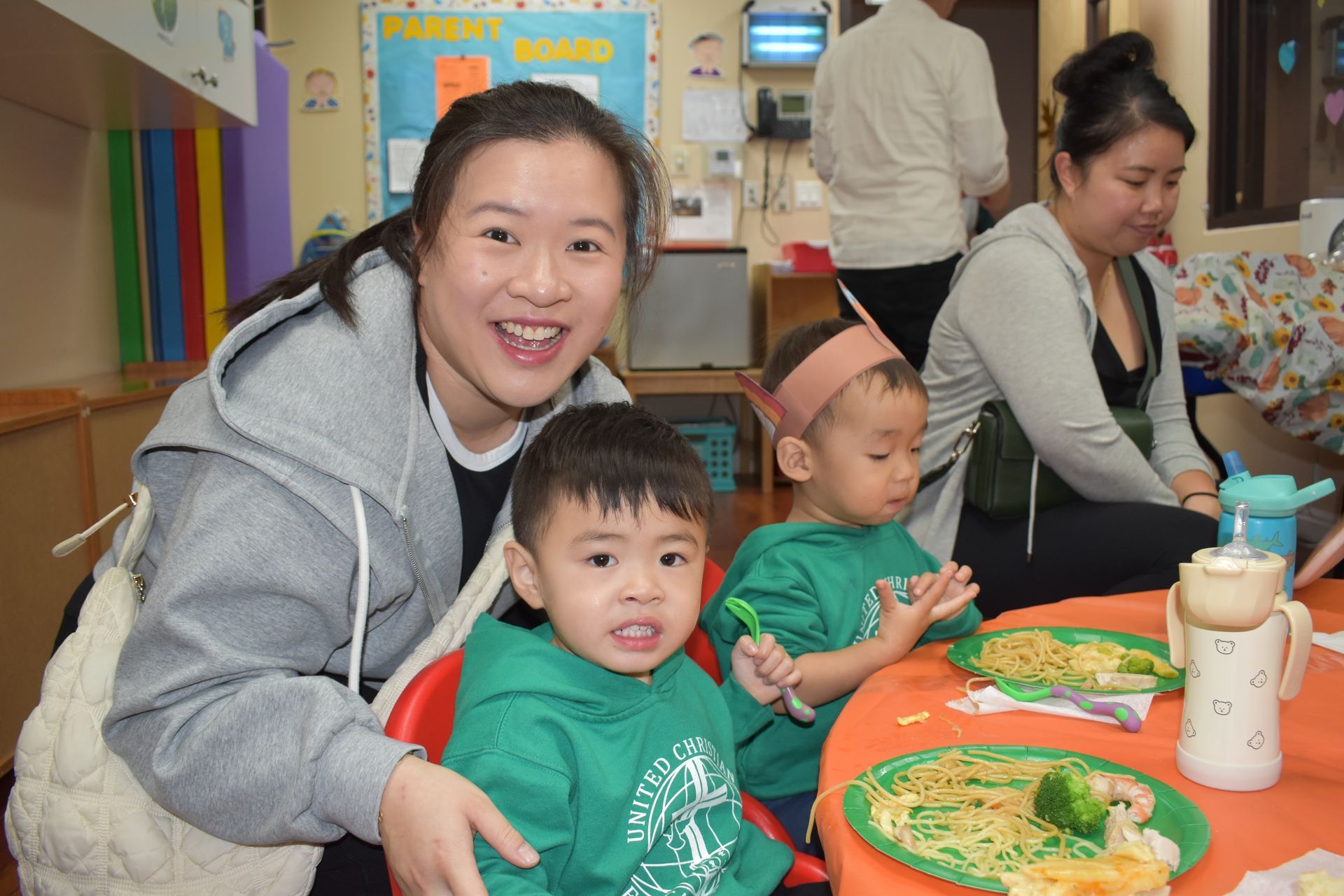 Woman smiles with two toddlers eating at a table; others in the background.