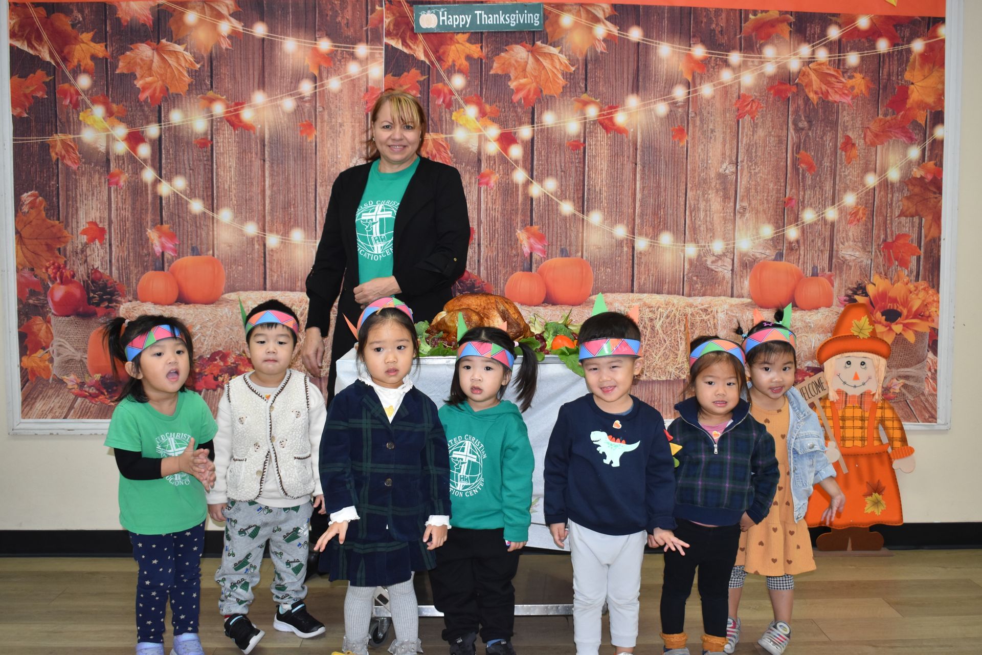 Children and teacher pose in front of a Thanksgiving backdrop, wearing festive headbands and outfits.