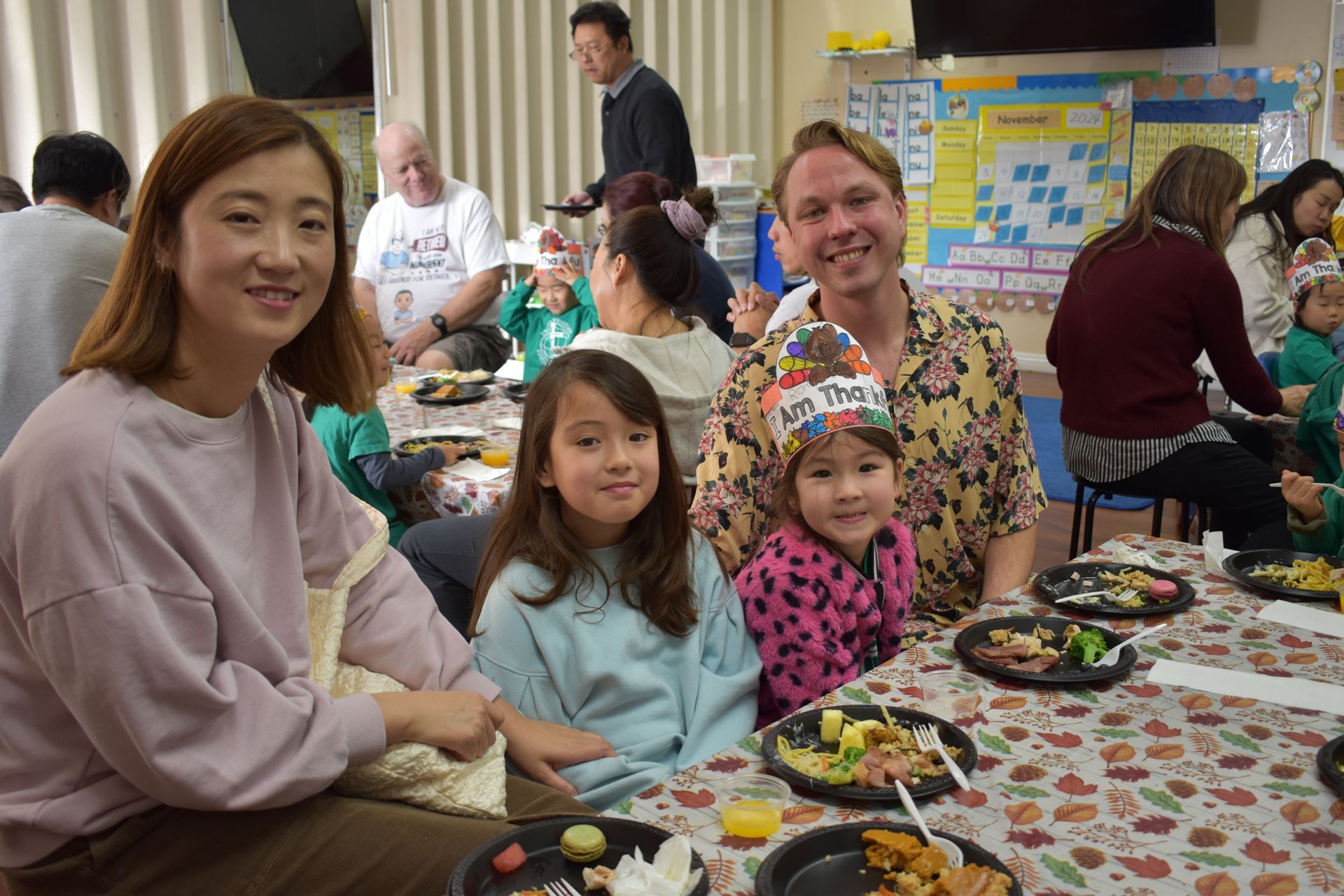 Family seated at a table with plates of food, smiling at the camera. Other people are in the background.
