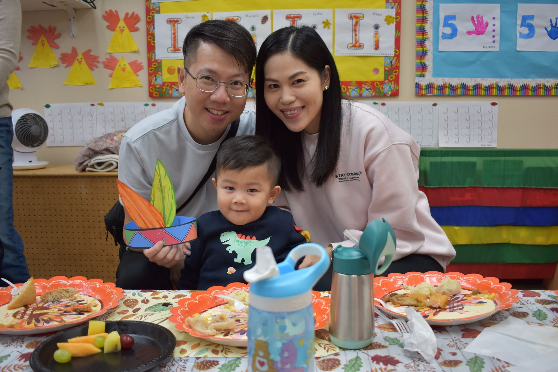 Family at a table smiling. Child holds handmade paper headwear. Plates of food and water bottles are visible.