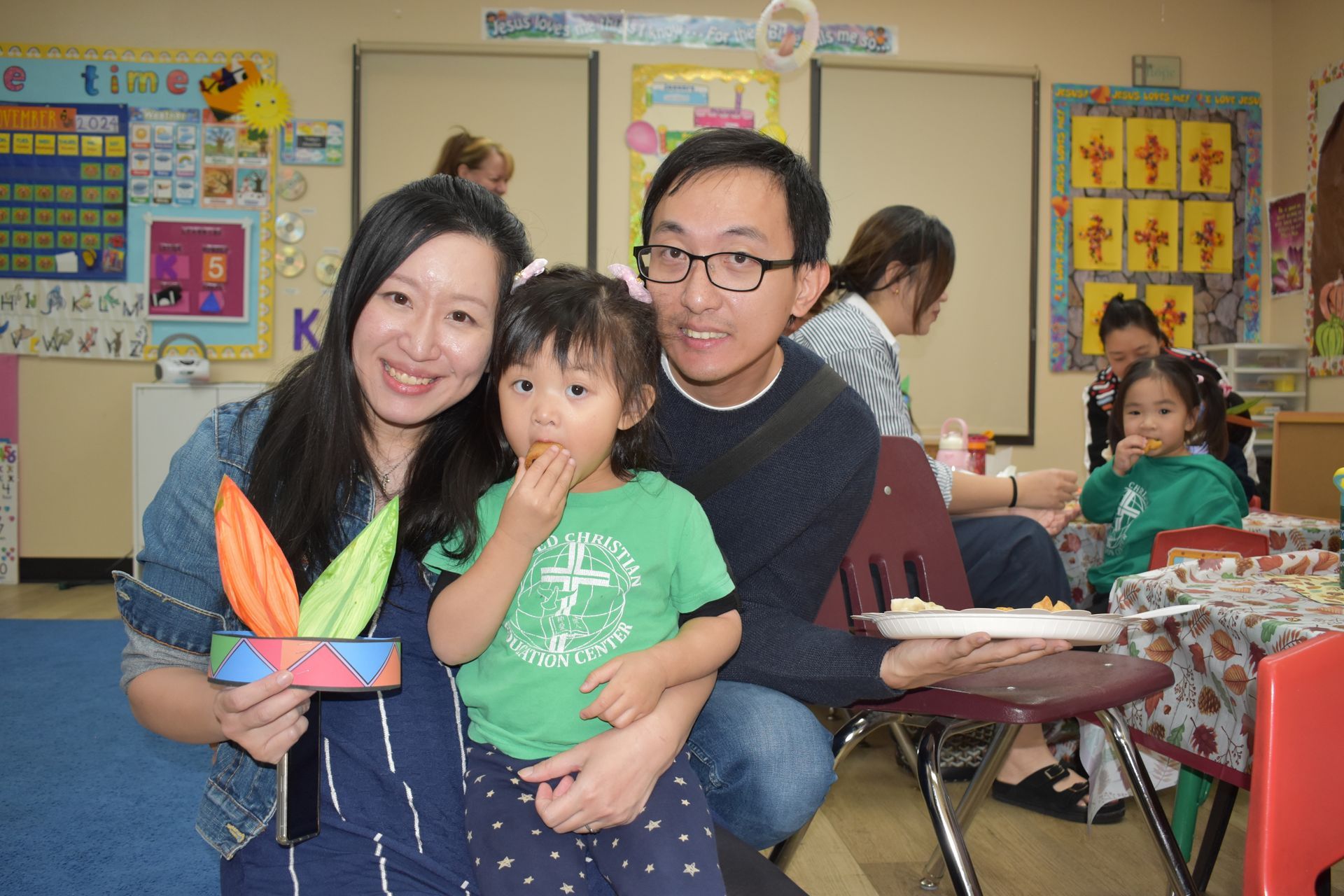 Family in classroom. Woman holds craft, child eats snack, man smiles. Classroom setting, other people present.