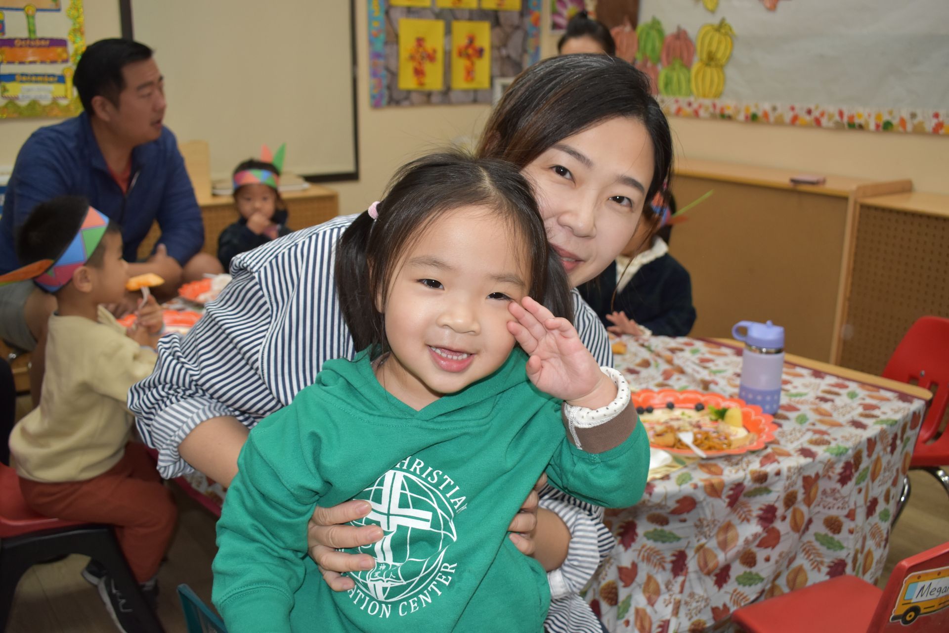 Woman and young child smiling, child saluting, in classroom with others, festive decorations.