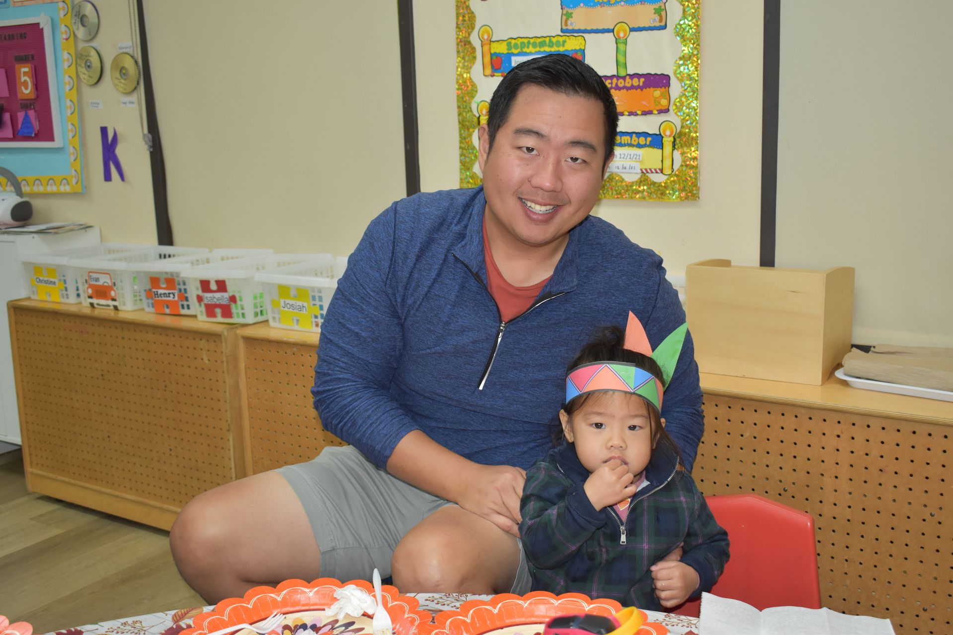 Man and child seated together, both wearing festive headbands, smiling in a classroom.