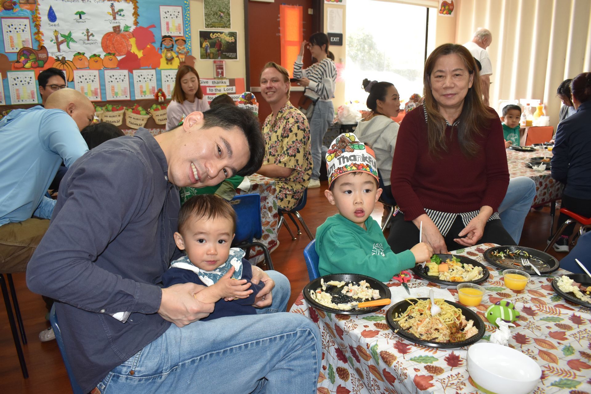 People at a table with food, including two children. The room has decorations and natural light.