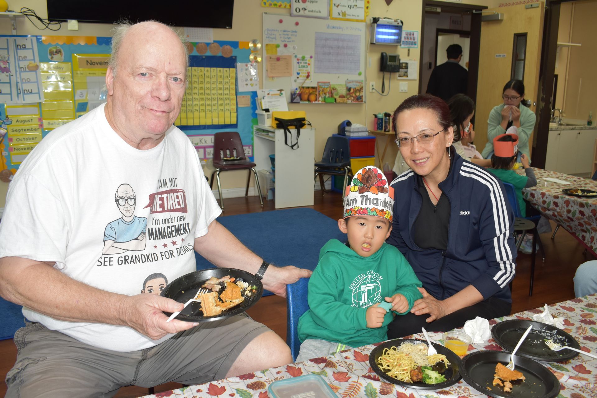 Three people at a table eating: an older man, a child wearing a party hat, and an adult woman. They are indoors.