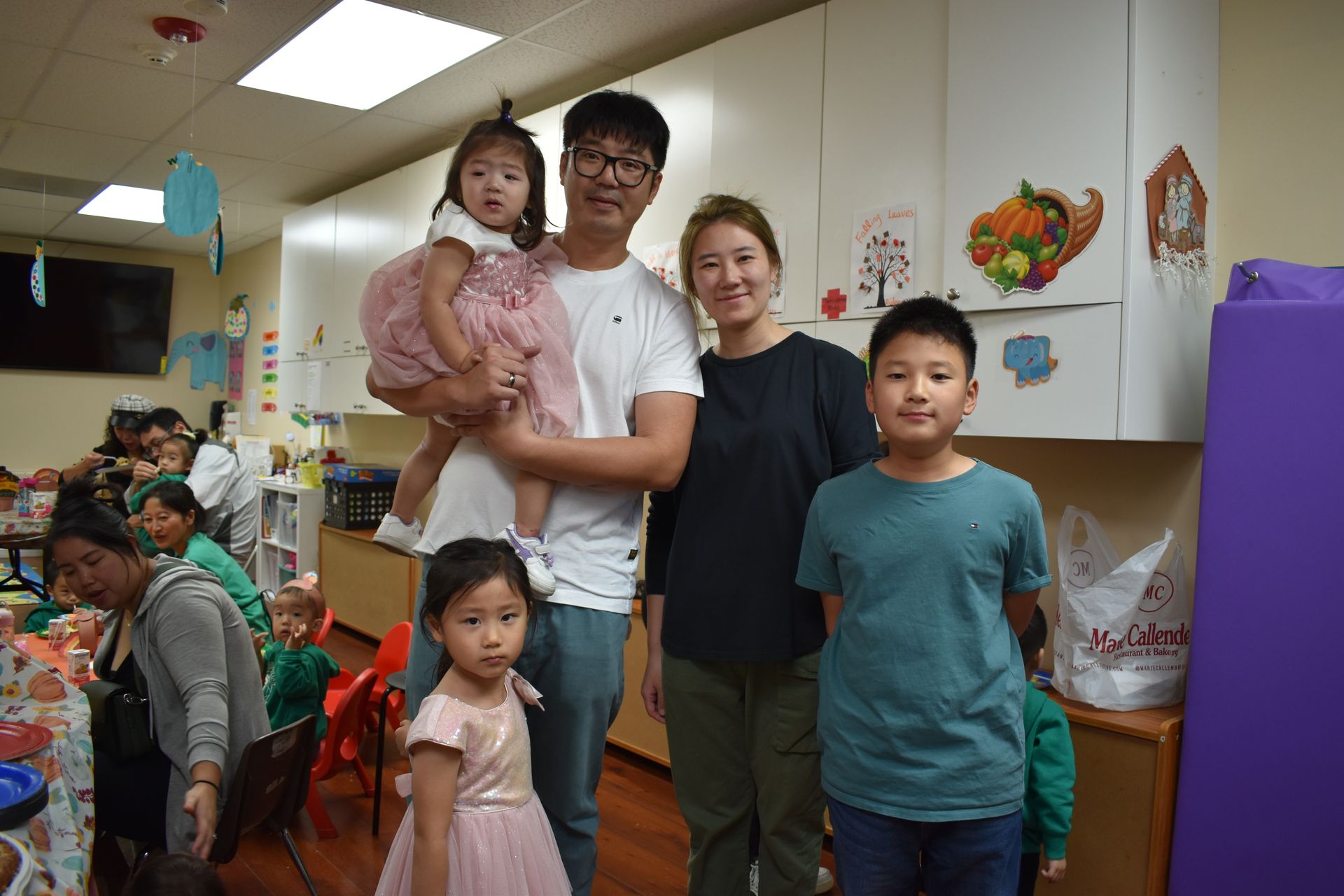 Family posing in a classroom; a man holds a child in a pink dress. A girl and boy stand in front.