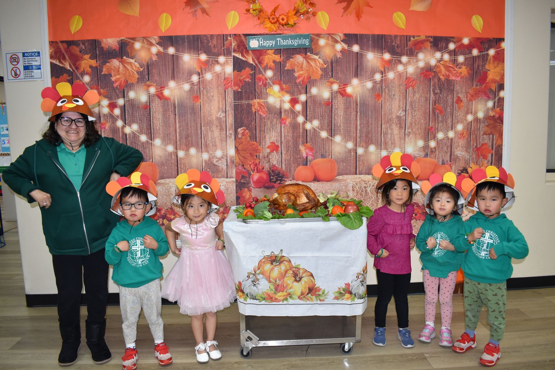 Teacher and children in turkey hats pose by a Thanksgiving display with food, pumpkins, and autumn decor.