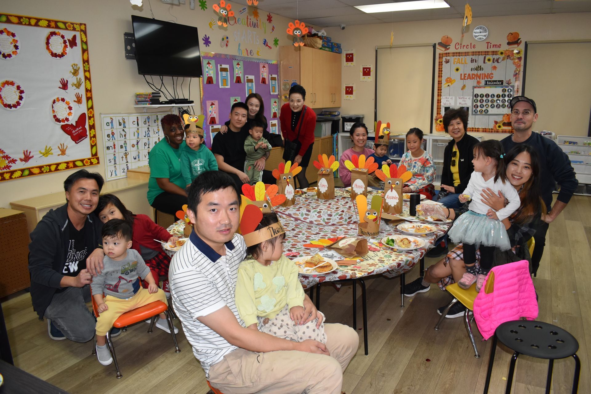 Group of people around a table set for Thanksgiving, children and adults smiling in a classroom decorated for the holiday.