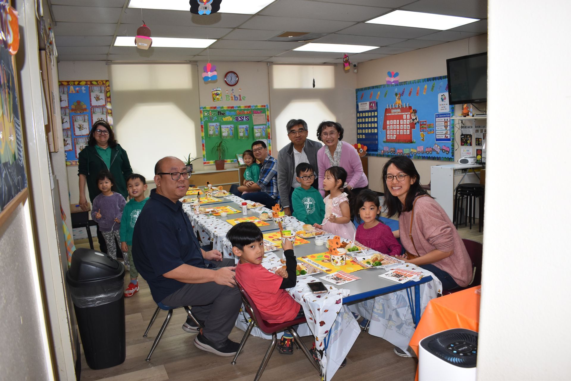 People seated at a decorated table in a classroom, with children and adults smiling.
