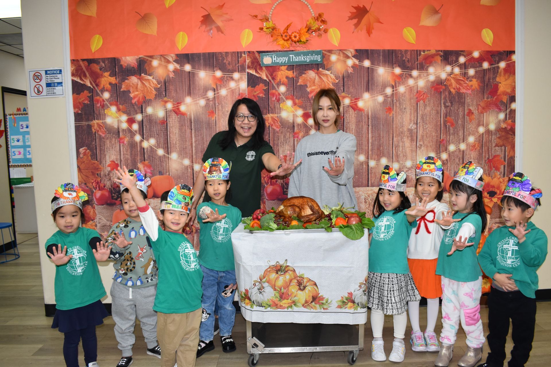 Children with teachers around Thanksgiving turkey display. Everyone smiles and poses, fall backdrop.