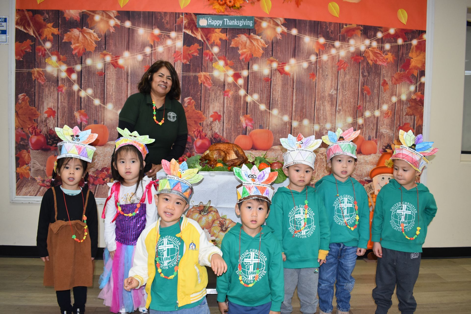 Children with Thanksgiving crafts and teacher pose in front of a festive backdrop.