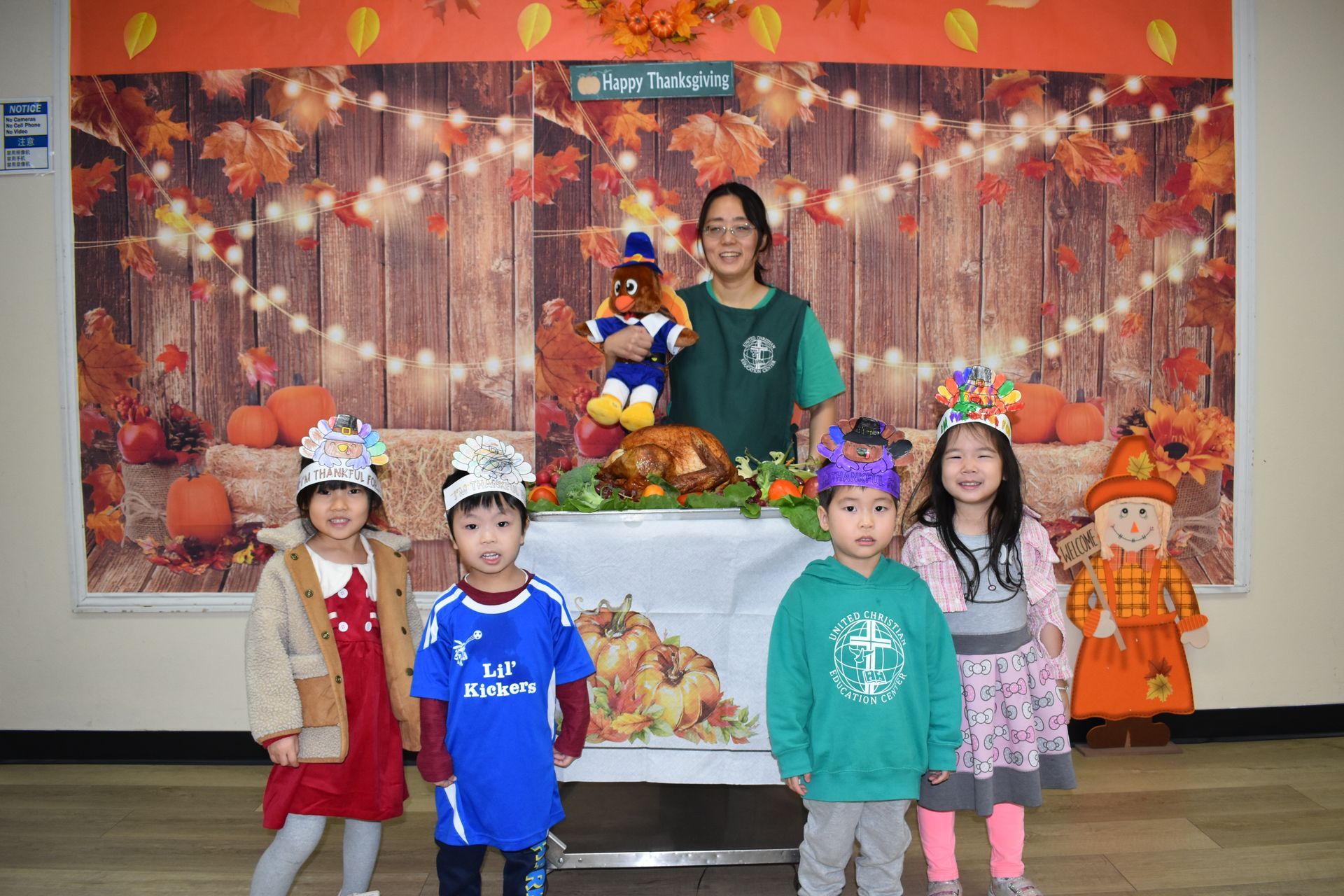 Group of children and adult with Thanksgiving display; pumpkins, turkey, and fall backdrop.