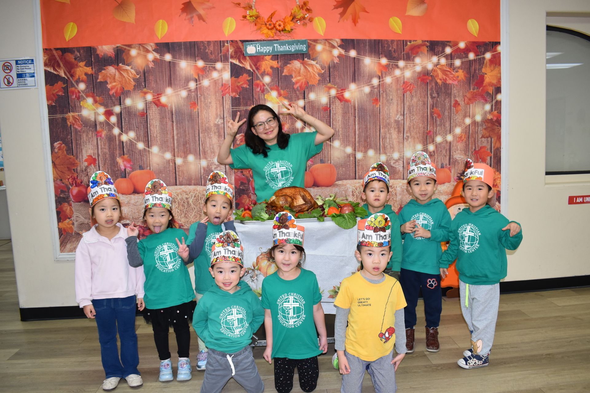 Group of children and a teacher in green shirts and turkey hats posing in front of a Thanksgiving backdrop.