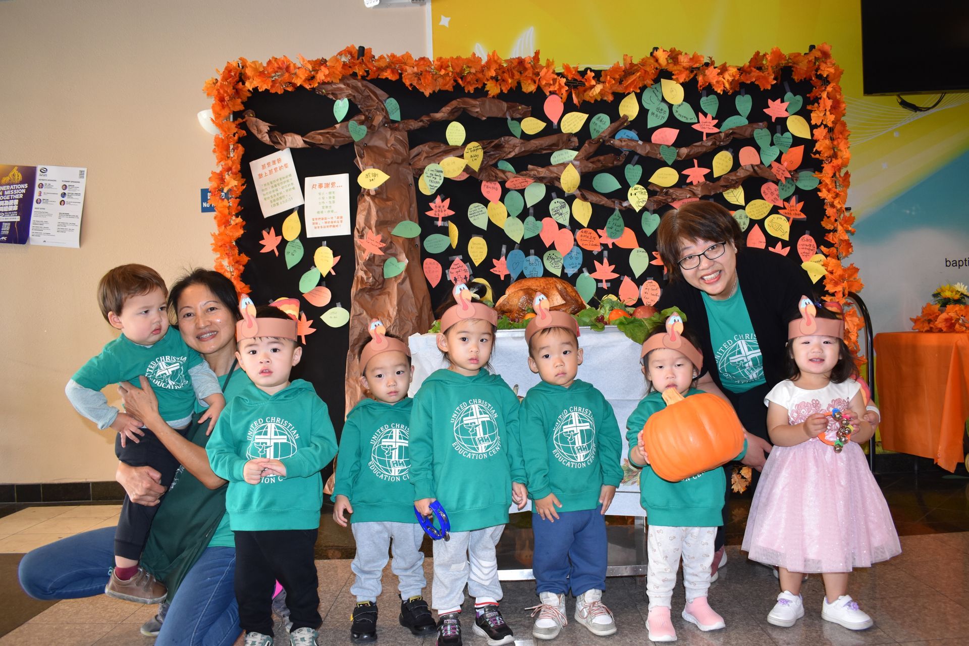 Children in green shirts and pumpkin hats pose with adults near a fall-themed backdrop.