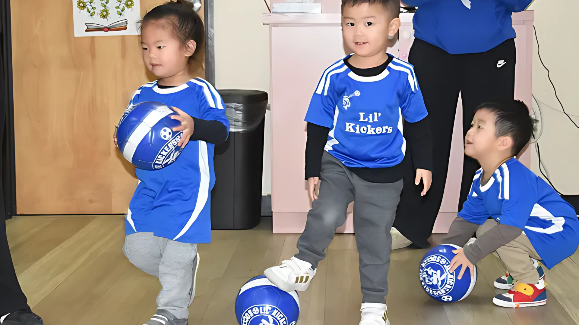 Three children in blue soccer jerseys playing with soccer balls indoors.