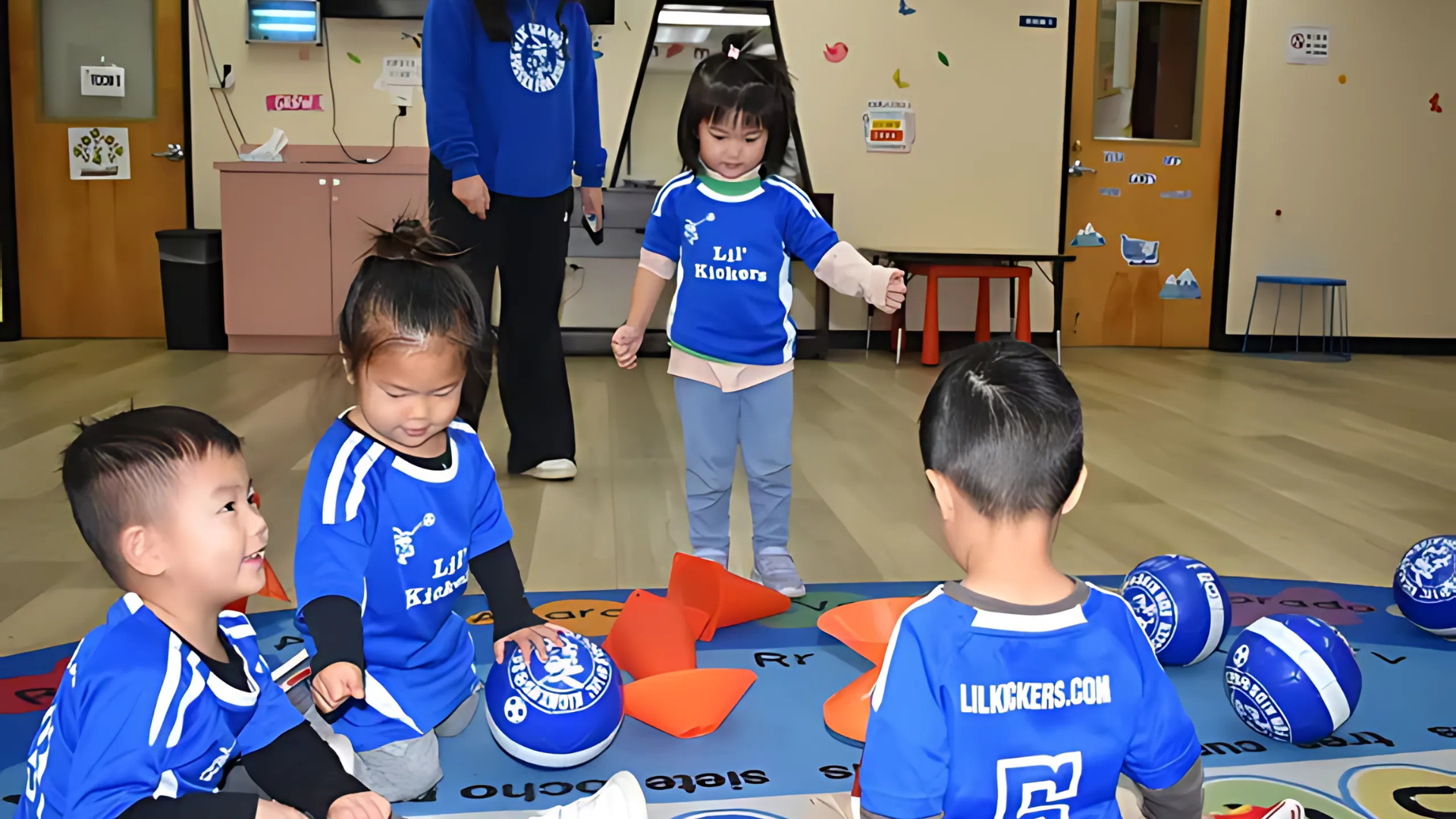Children in blue shirts playing with balls and orange cones on a blue mat, indoors.