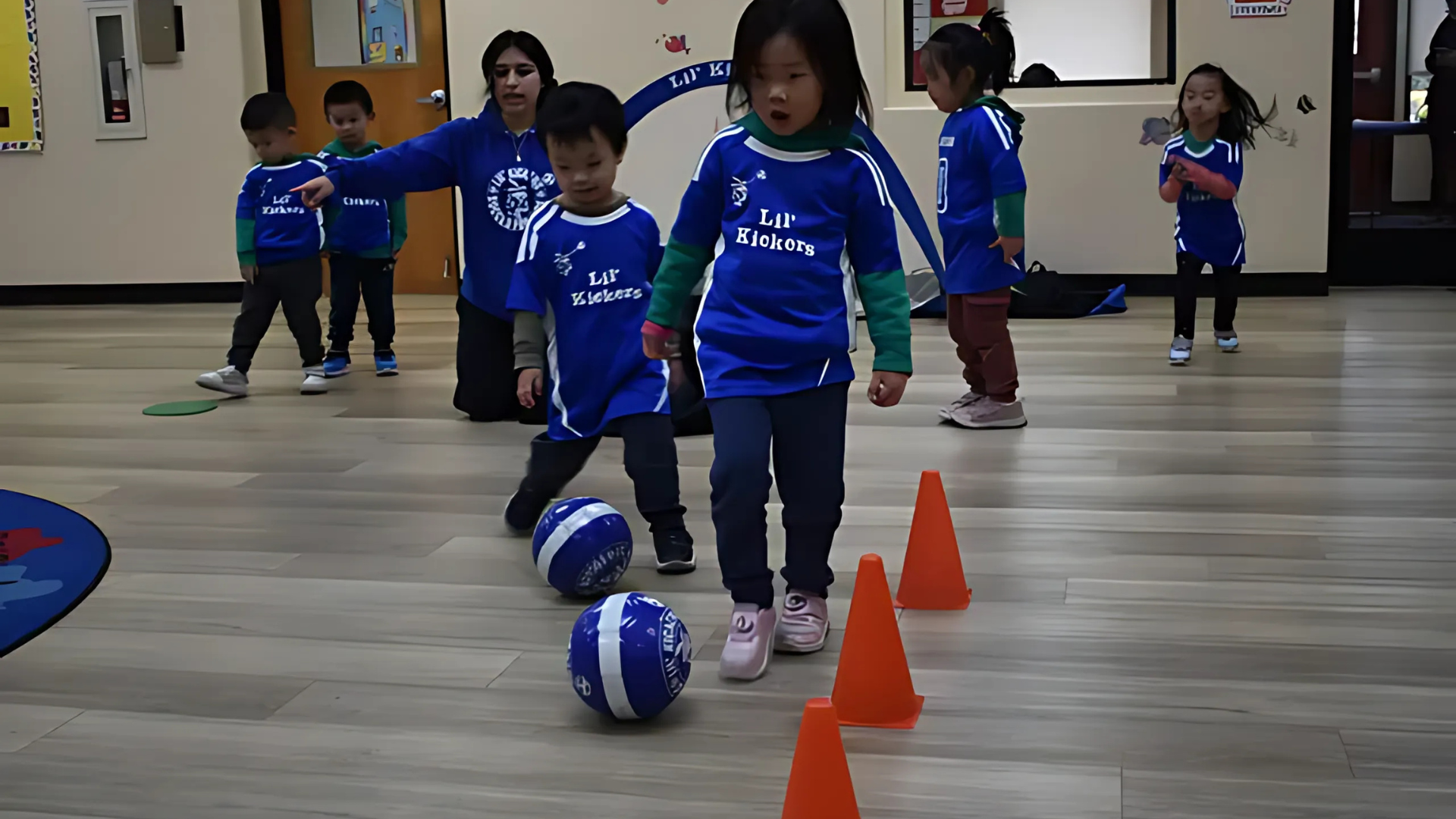 Children in blue jerseys play soccer indoors with cones and an instructor.