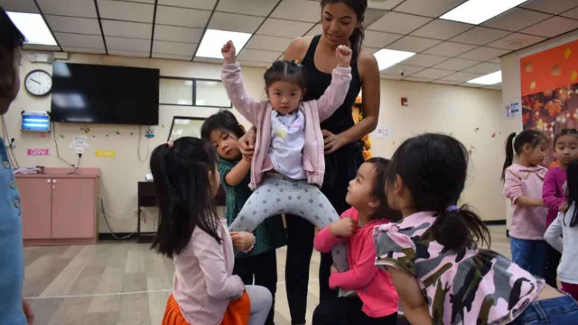 Children and an adult support a child who is jumping in a classroom.