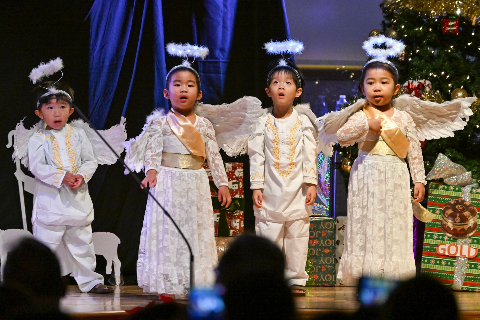 Four children in angel costumes sing on stage with Christmas tree in background.